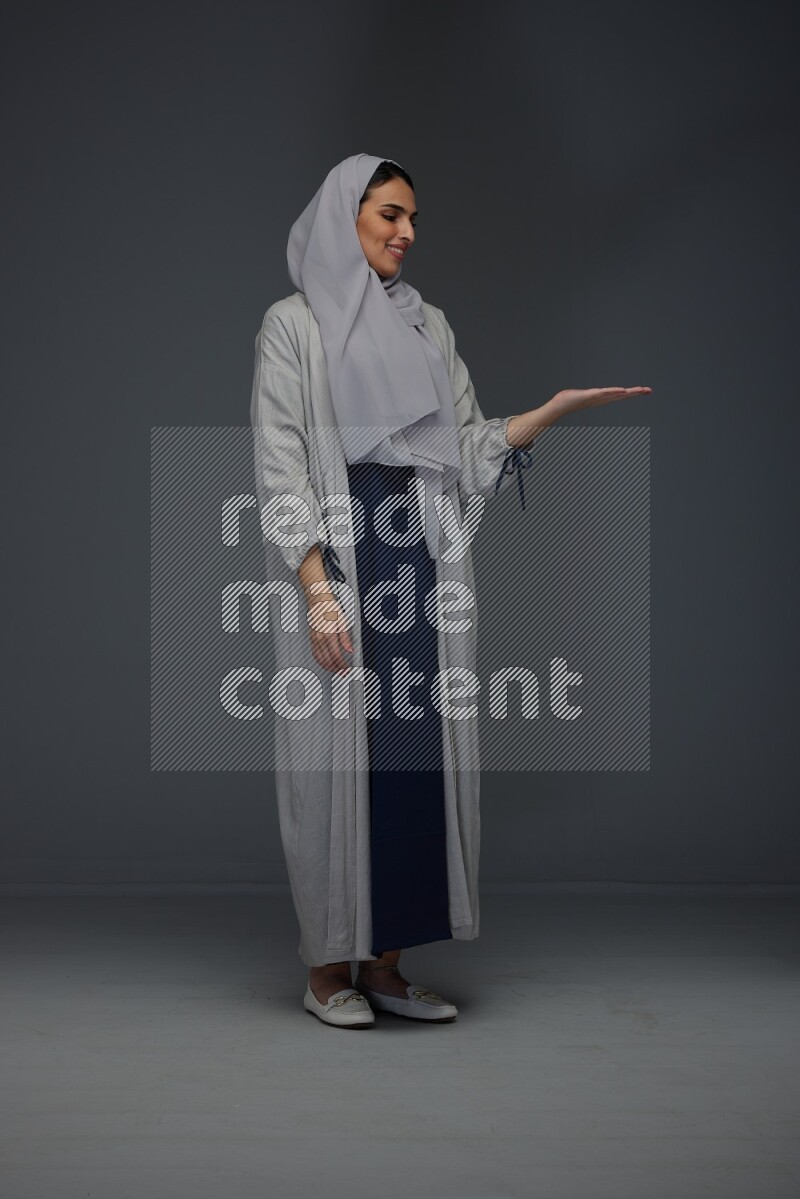 A Saudi woman wearing a light gray Abaya and head scarf standing and pointing in different directions eye level on a grey background
