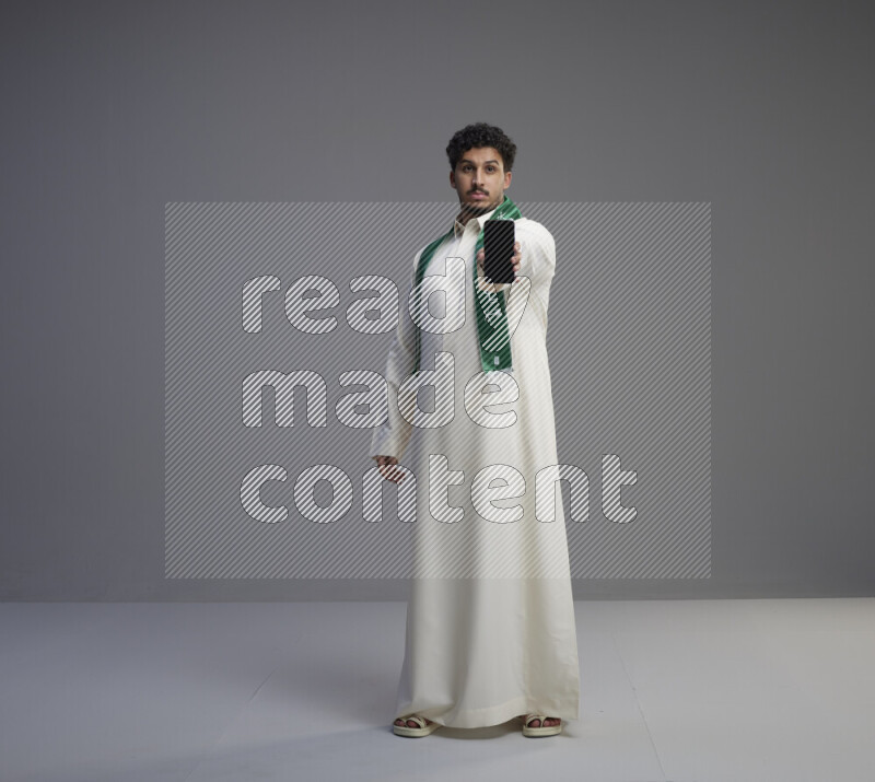 A Saudi man standing wearing thob and Saudi flag scarf showing phone to camera on gray background