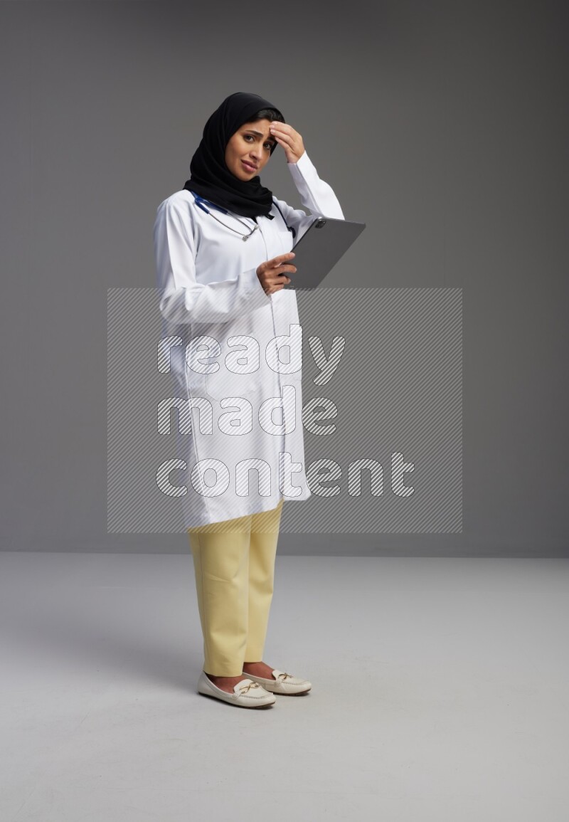 Saudi woman wearing lab coat with stethoscope standing working on tablet on Gray background