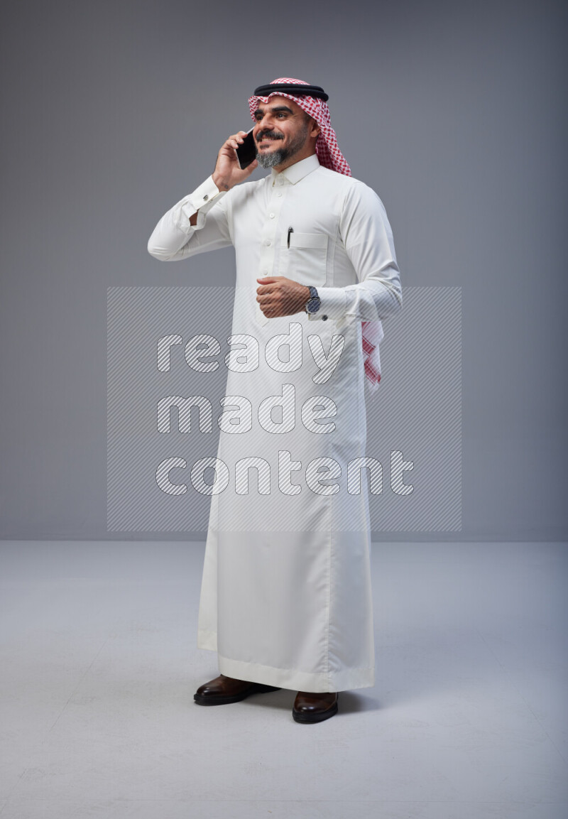 Saudi man Wearing Thob and red Shomag standing talking on phone on Gray background