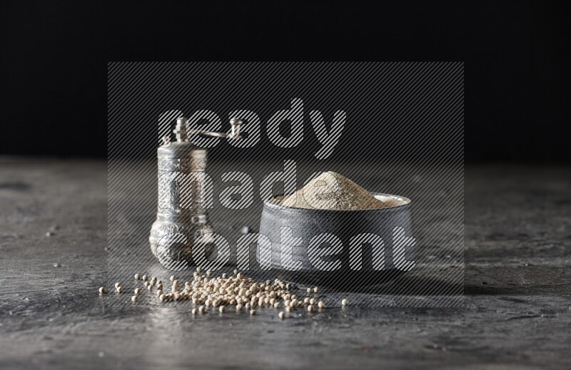 Black pottery bowl full of white pepper powder with pepper beads and metal pepper grinder on textured black flooring