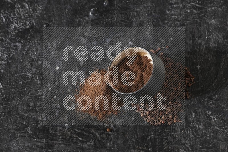 A black pottery bowl full of cloves powder spilled out of it with whole grains on a textured black flooring