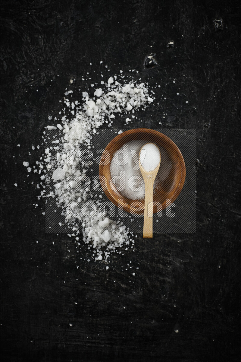 A pottery plate full of fine salt with bunch of coarse salt beside it on black background