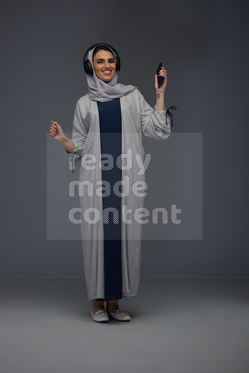 A Saudi woman wearing a light gray Abaya and head scarf standing and listening to music on a grey background