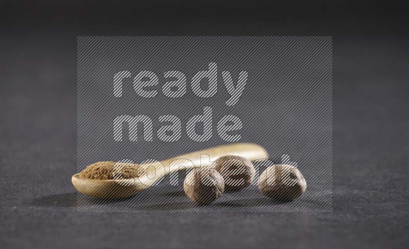 A wooden spoon full of nutmeg powder with whole nutmeg seeds beside it on a black flooring