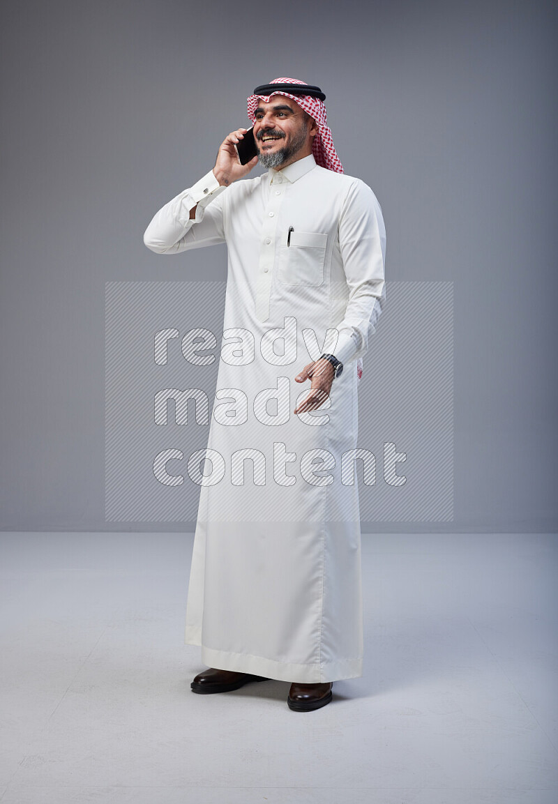 Saudi man Wearing Thob and red Shomag standing talking on phone on Gray background