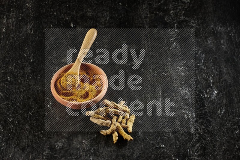 A wooden bowl and a wooden spoon full of turmeric powder with dried turmeric fingers on textured black flooring