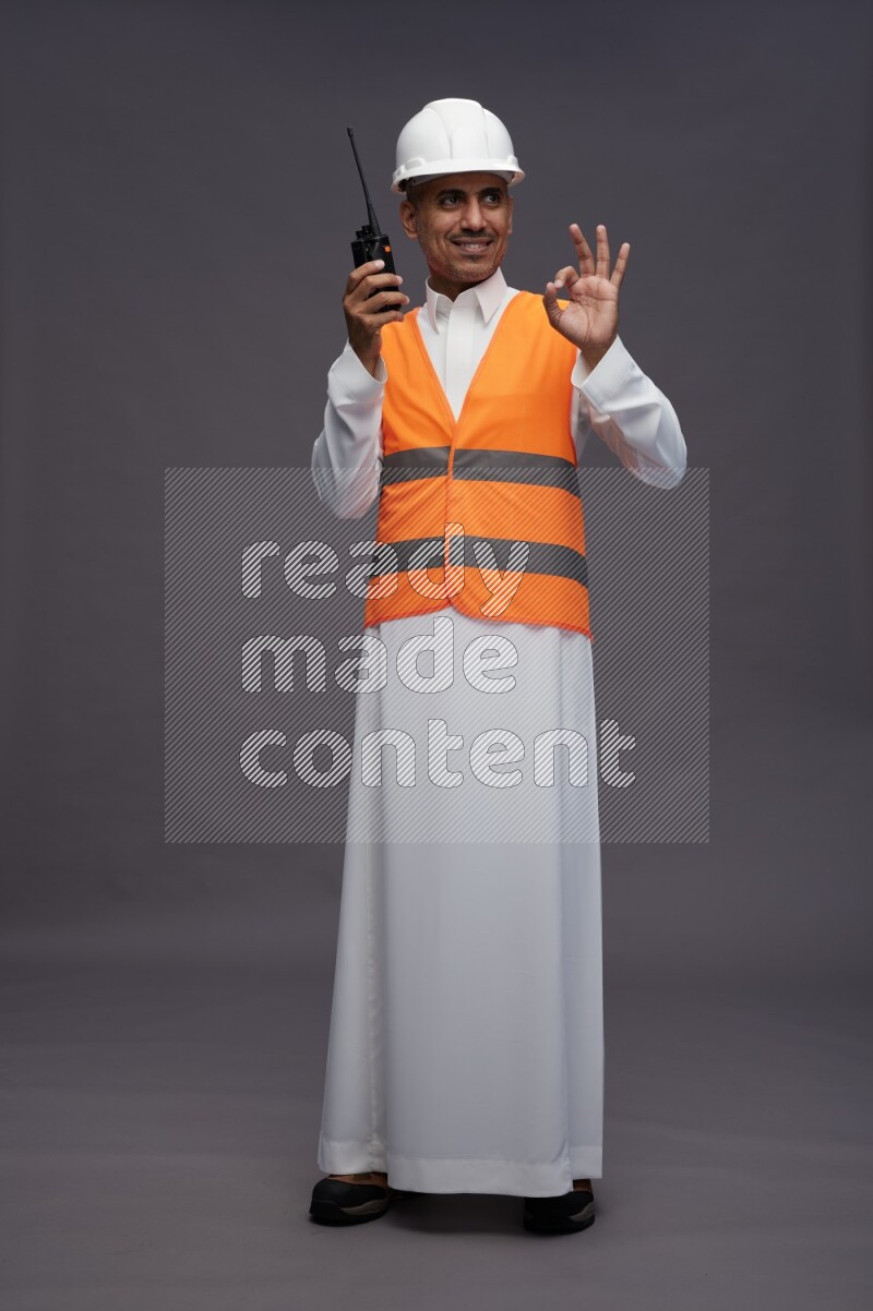 Saudi man wearing thob with engineer vest standing holding walkie-talkie on gray background
