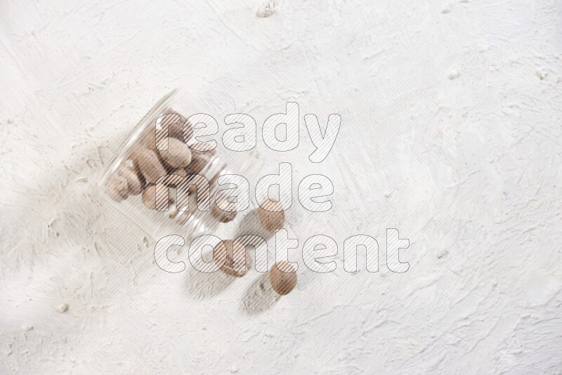 A glass jar full of nutmeg flipped and the seeds spread out on a textured white flooring