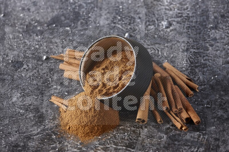 Black pottery bowl over filled with cinnamon powder and cinnamon sticks around the bowl on a textured black background