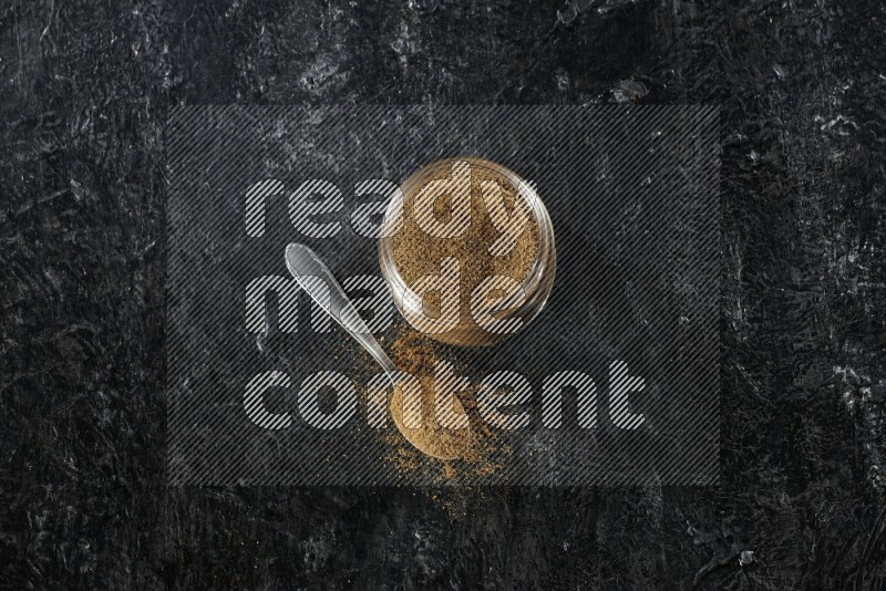 وعاء زجاجي للتوابل وملعقة معدنية ممتلئين ببودرة الكمون على خلفية سوداء