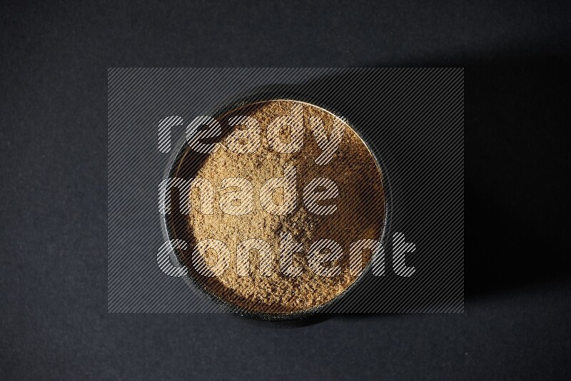 A black pottery bowl full of cumin powder on a black flooring