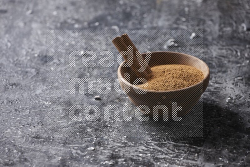 Wooden bowl full of cinnamon powder and a cinnamon stick on a textured black background