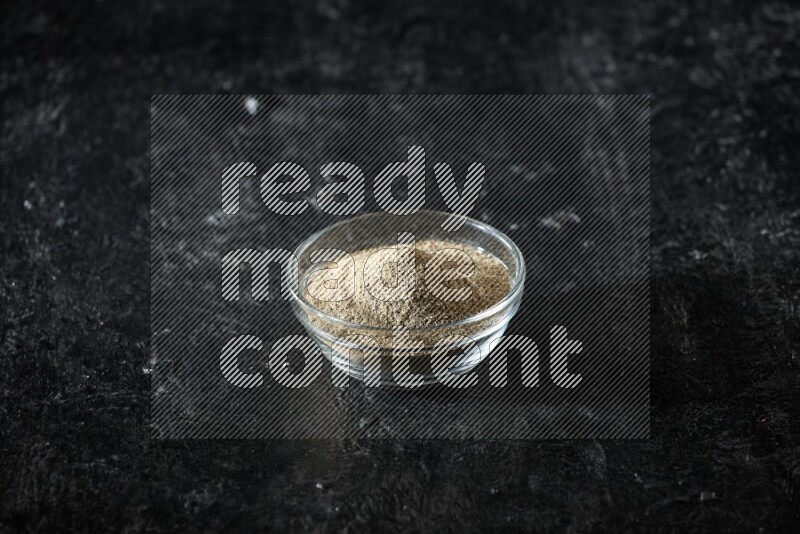 A glass bowl full of cardamom powder on textured black flooring