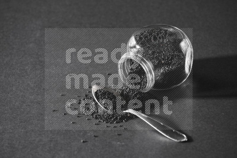 A flipped glass spice jar full of black seeds and the seeds spread out with a metal spoon full it on a black flooring