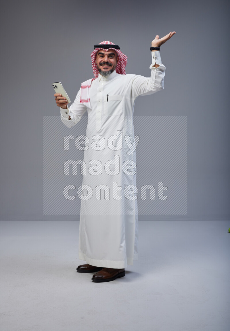 Saudi man Wearing Thob and red Shomag standing texting on phone on Gray background
