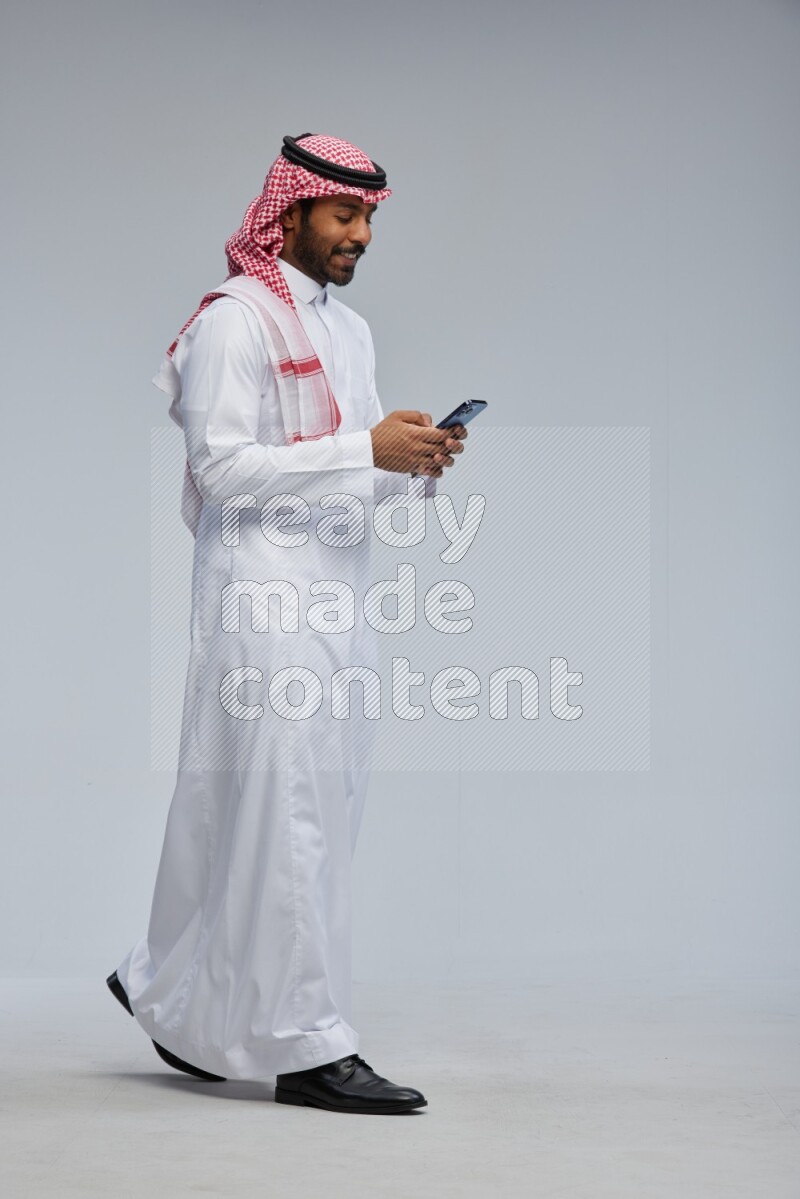 Saudi man Wearing Thob and shomag standing texting on phone on Gray background