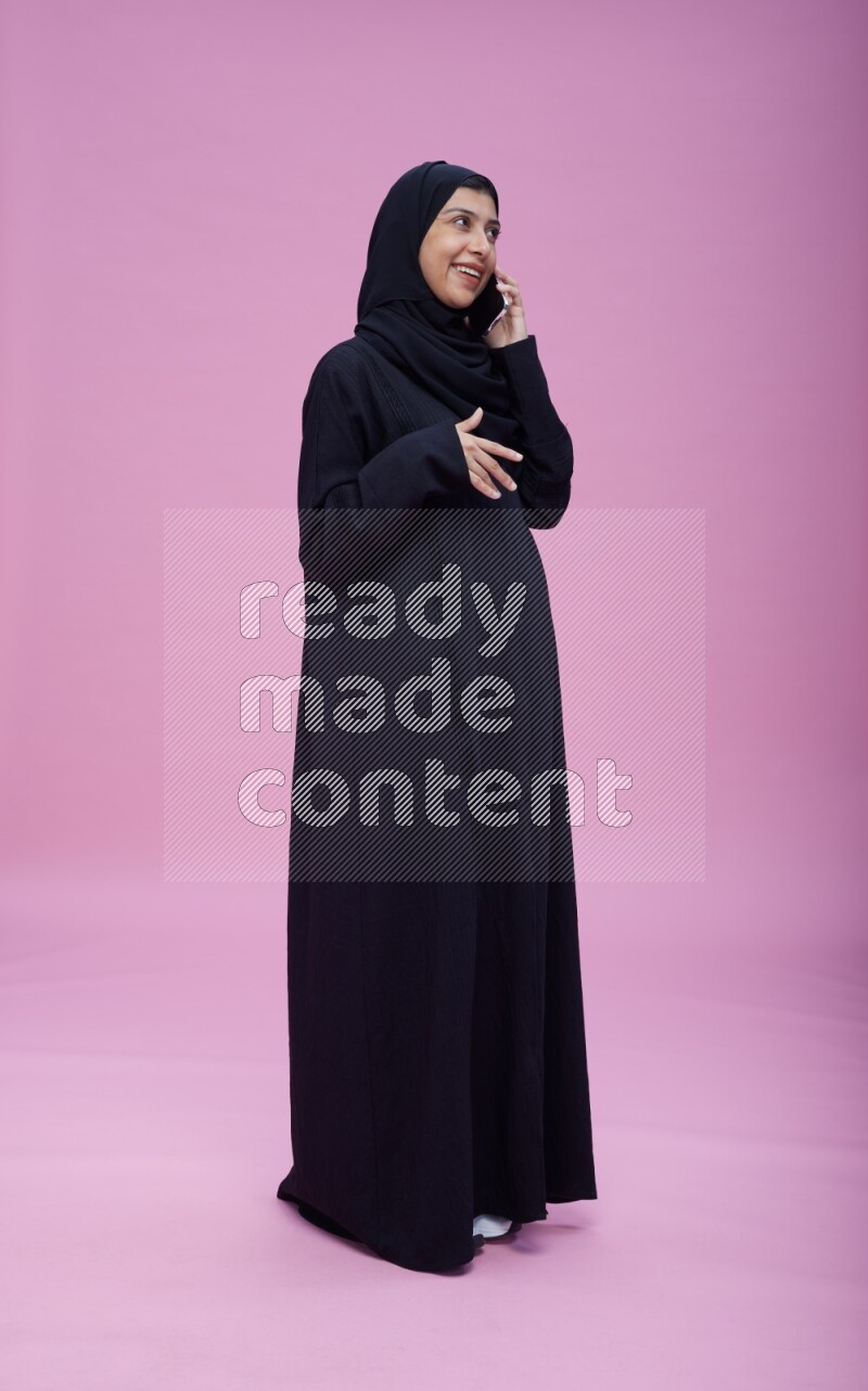 Saudi woman wearing Abaya standing talking on phone on pink background