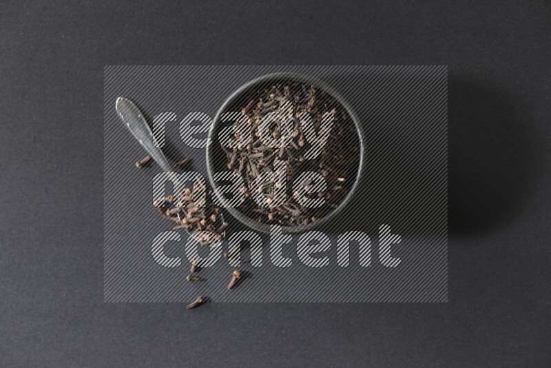 A black pottery bowl full of cloves and a metal spoon next to it on a black flooring
