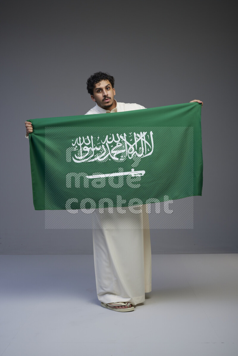 A Saudi man standing wearing thob holding big Saudi flag on gray background