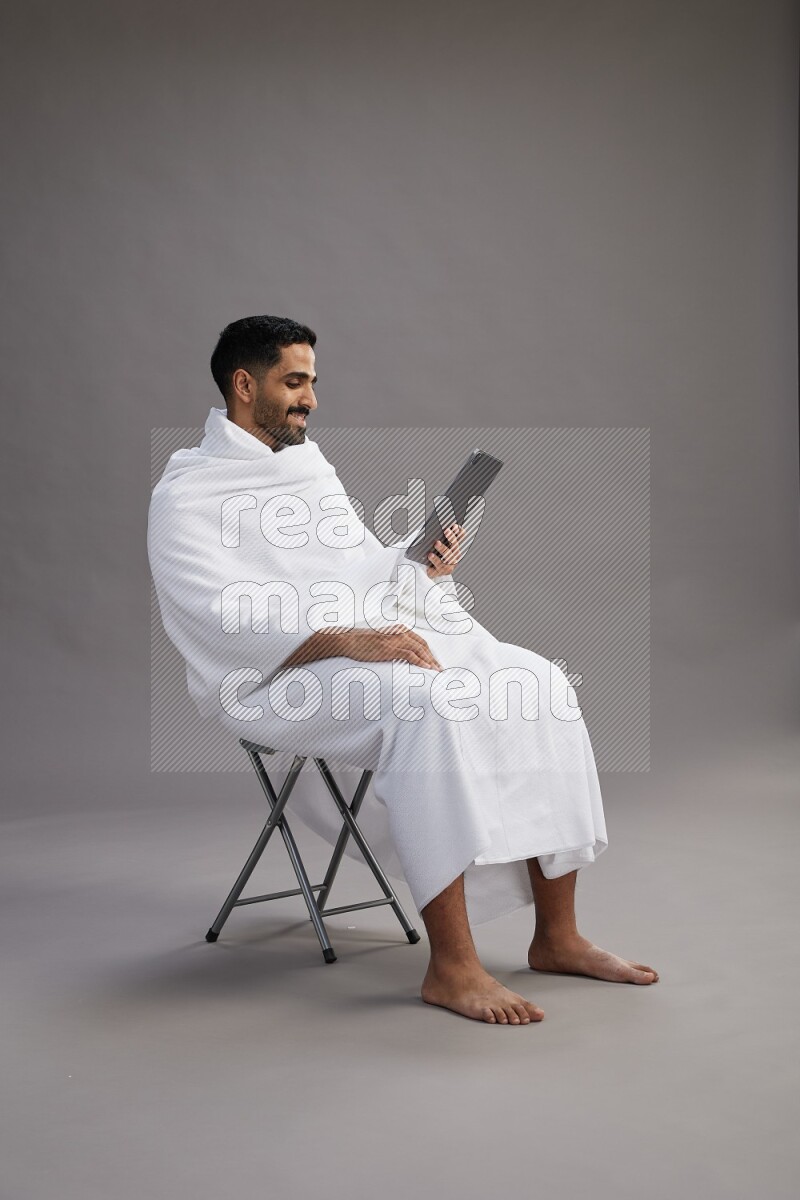 A man wearing Ehram sitting on chair holding a tablet on gray background