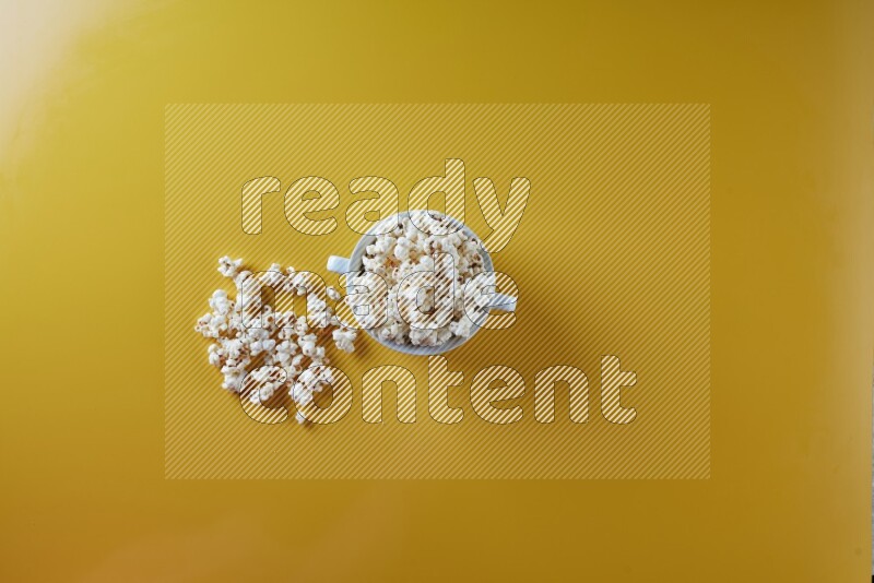 A white ceramic bowl full of popcorn with popcorn beside it on a yellow background in a top view shot