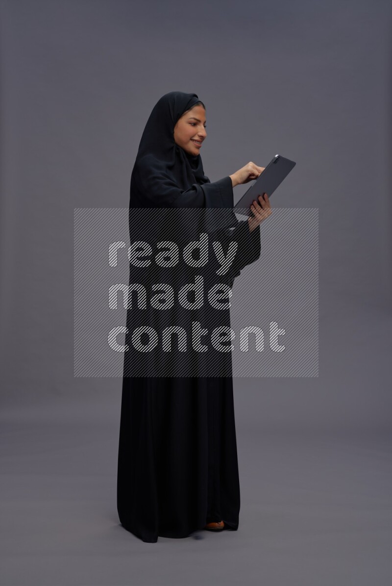Saudi woman wearing Abaya standing working on tablet on gray background