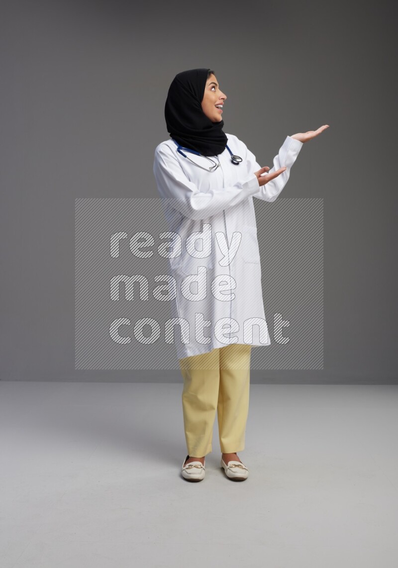 Saudi woman wearing lab coat with stethoscope standing interacting with the camera on Gray background