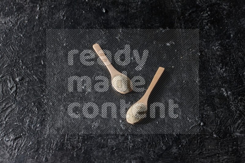 2 wooden spoons full of white pepper powder on textured black flooring