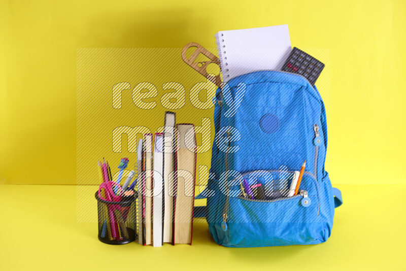A school bag with assorted school supplies in and beside it on yellow background