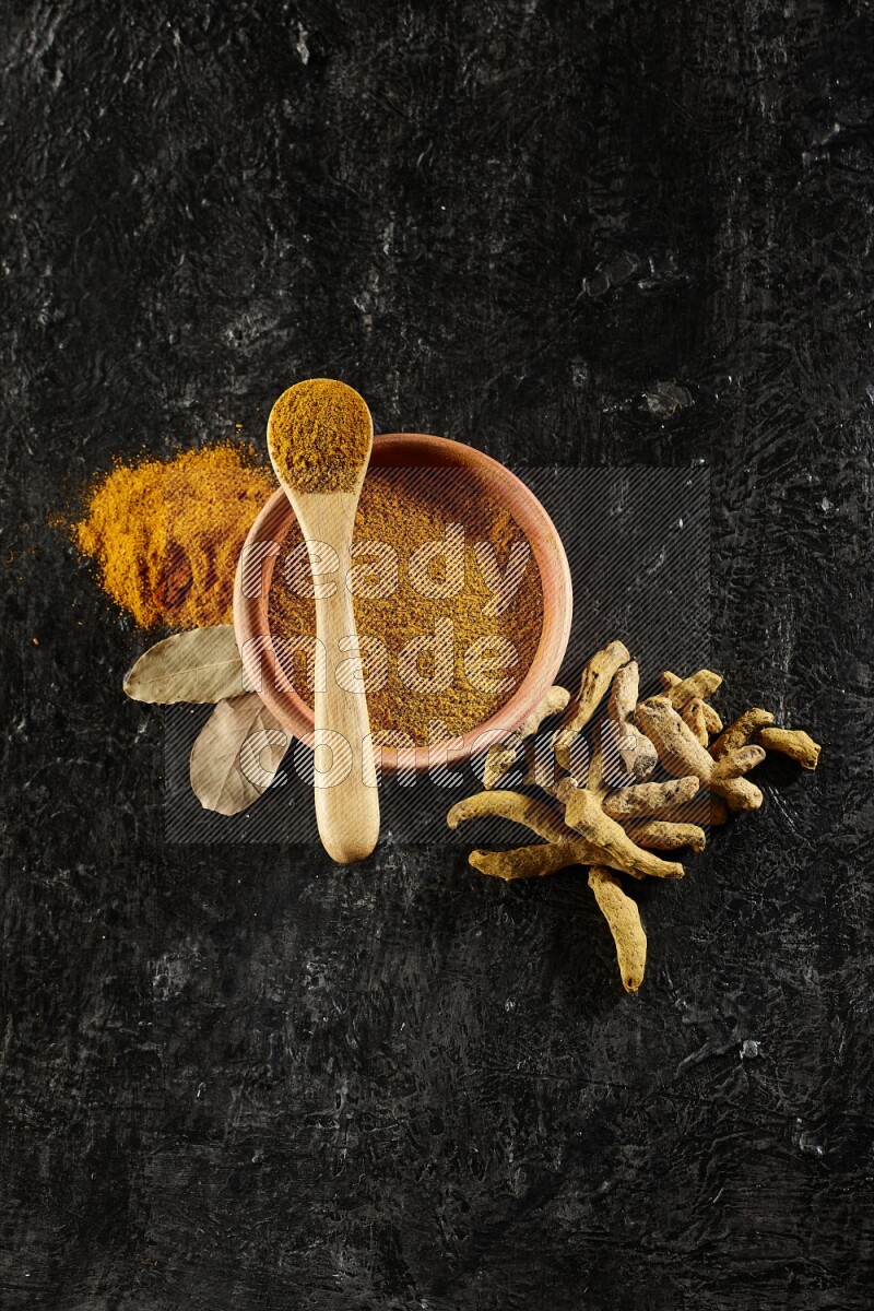 A wooden bowl and a wooden spoon full of turmeric powder with dried turmeric fingers on textured black flooring