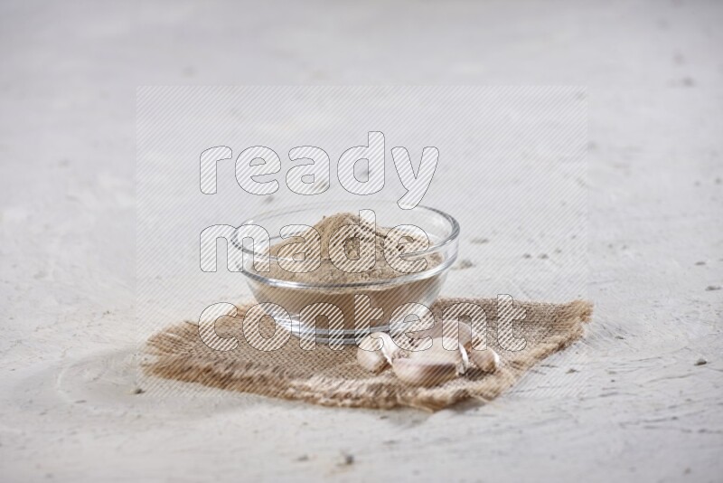 A glass bowl full of garlic powder placed on burlap fabric with garlic cloves on a textured white flooring