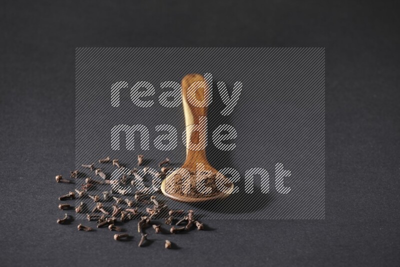 A wooden ladle full of cloves powder and some of whole cloves around it on a black flooring