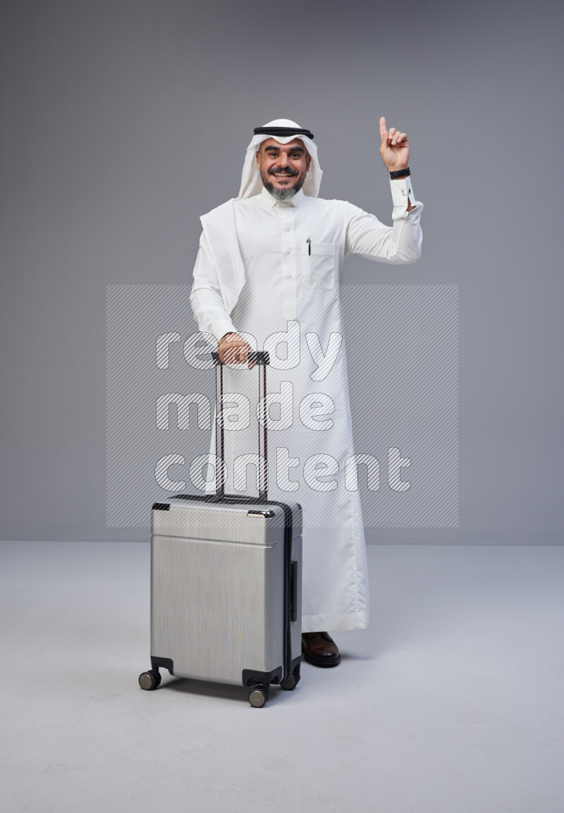 Saudi man wearing Thob and white Shomag standing holding Travel bag on Gray background