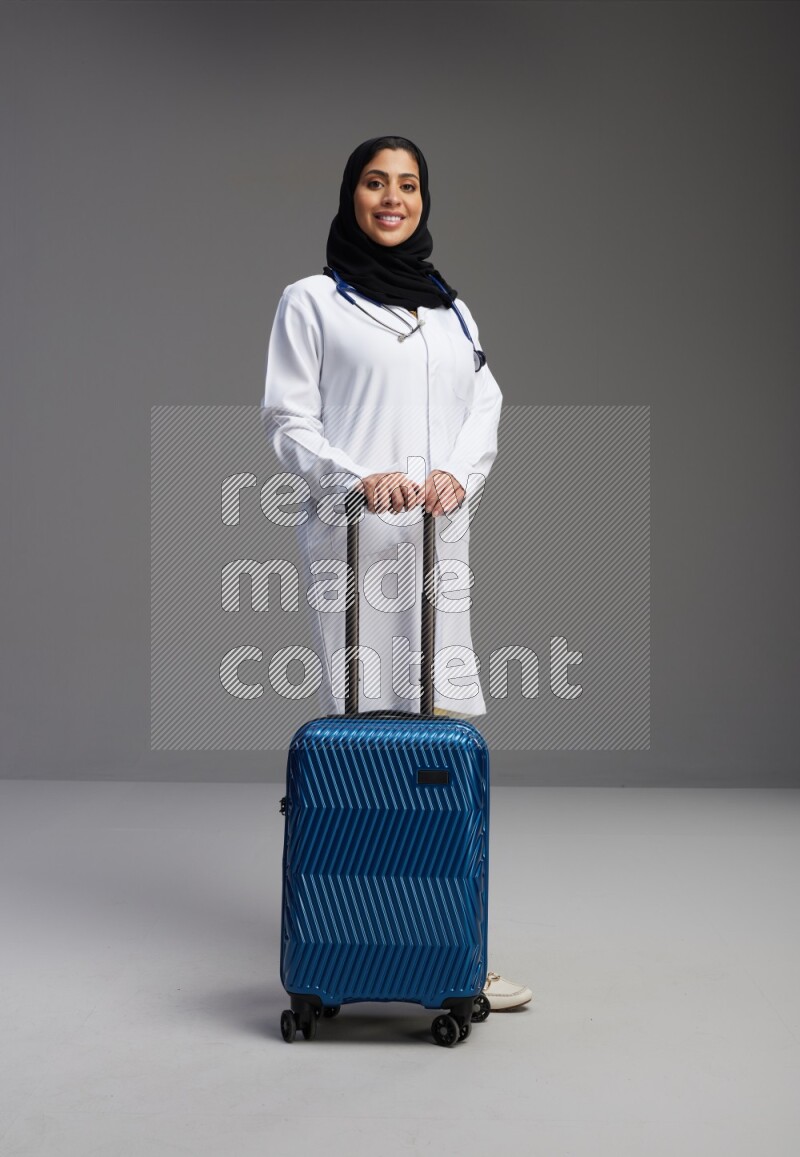 Saudi woman wearing lab coat with stethoscope standing holding Travel bag on Gray background