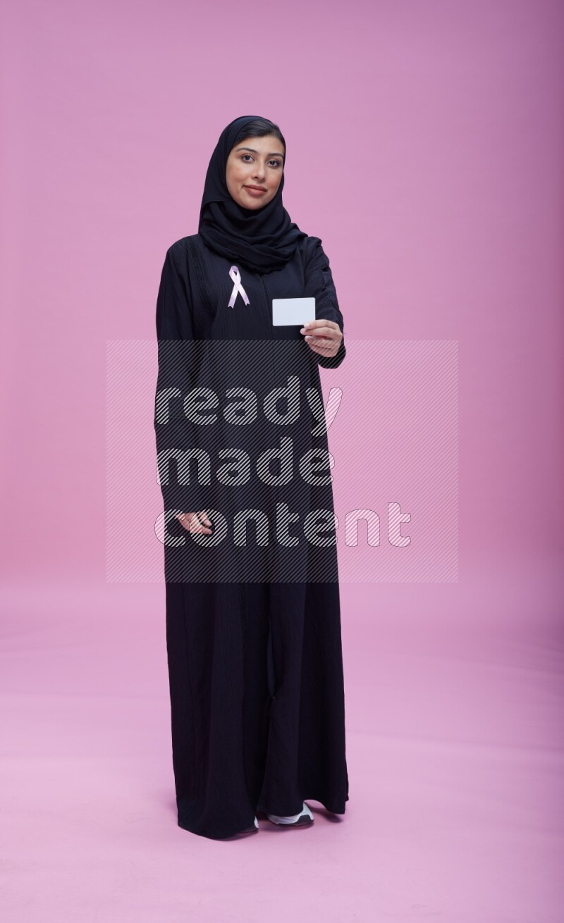 Saudi woman wearing pink ribbon on Abaya standing holding ATM card on pink background