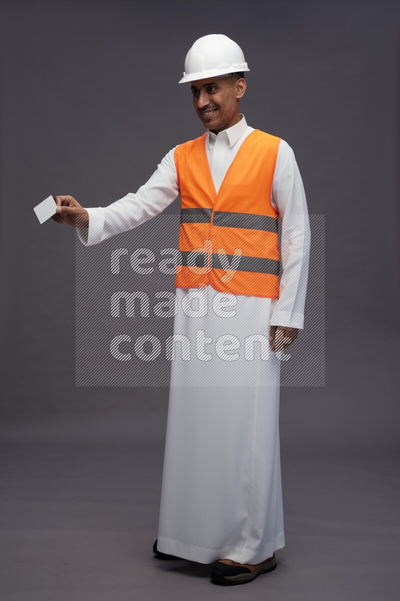 Saudi man wearing thob with engineer vest standing holding ATM card on gray background