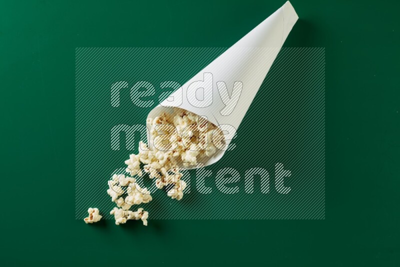 A paper cone full of popcorn and some popcorn came out of it on a green background in a top veiw shot