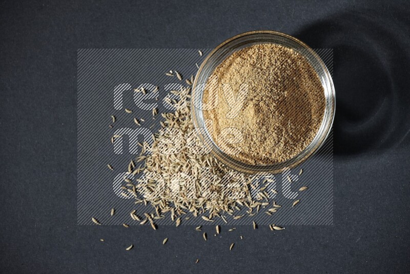 A glass bowl full of cumin powder with cumin seeds beside it on black flooring