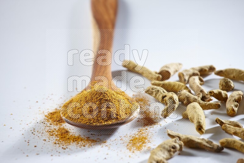 A wooden ladle full of turmeric powder and dried turmeric fingers beside it on white flooring