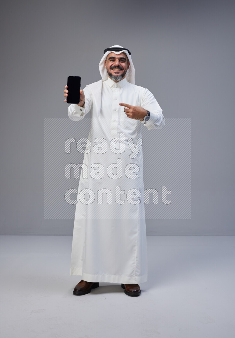 Saudi man Wearing Thob and white Shomag standing showing phone to camera on Gray background
