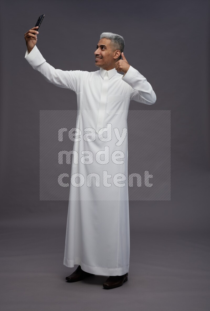 Saudi man wearing thob standing taking selfie on gray background