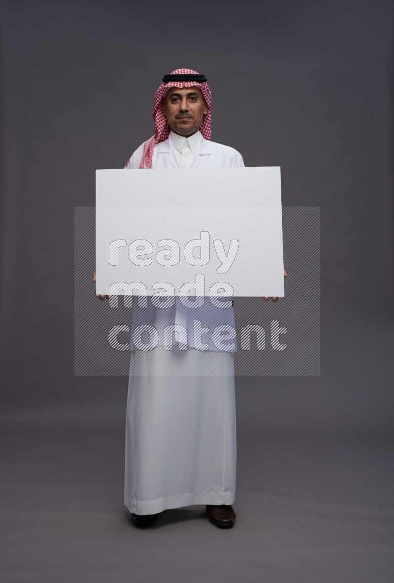 Saudi man wearing thob with lab coat and shomag with pocket employee badge standing holding board on gray background