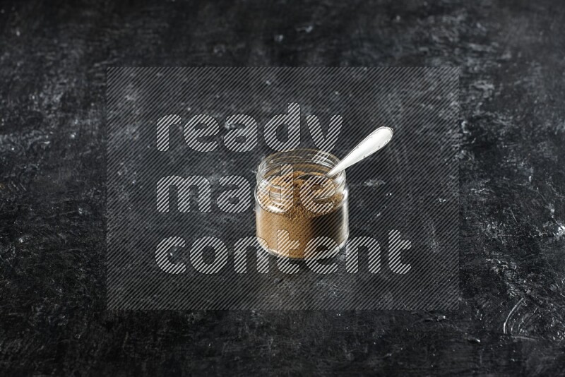 A glass jar and a metal spoon full of cumin powder on a textured black flooring