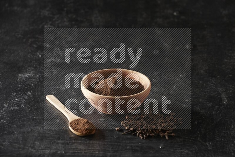 A wooden bowl and a wooden spoon full of cloves powder with spreaded cloves on a textured black flooring