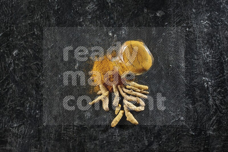 A flipped glass spice jar full of turmeric powder and powder spilled out of it with dried whole fingers on textured black flooring