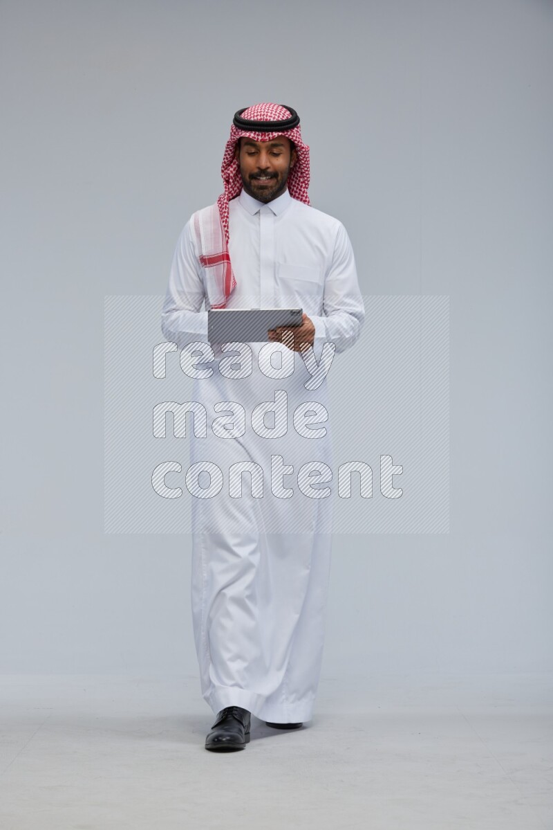 Saudi man Wearing Thob and shomag standing working on tablet on Gray background