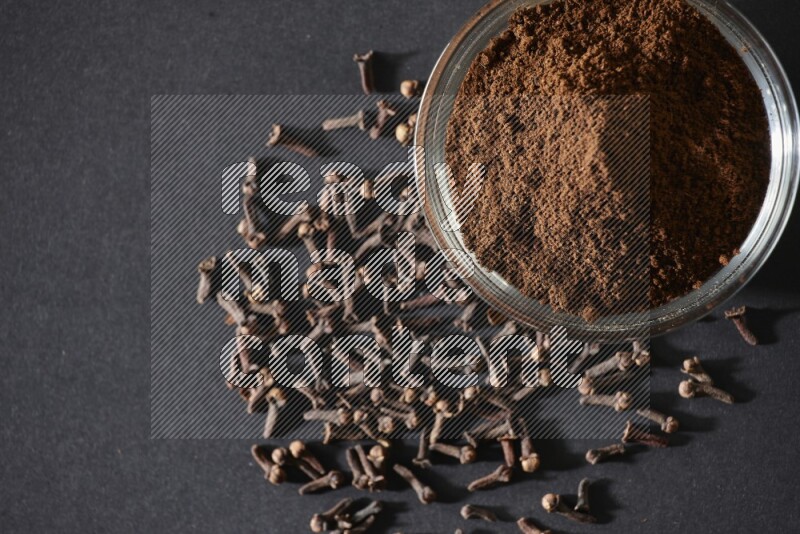 A glass bowl full of cloves powder with cloves grains spread on black flooring
