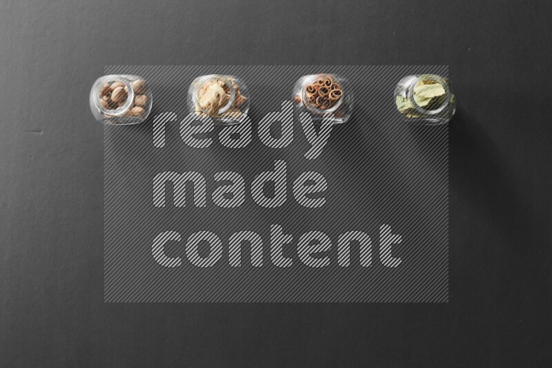 4 glass spice jars full of laurel bay leaves, ginger, cinnamon and nutmeg on black flooring