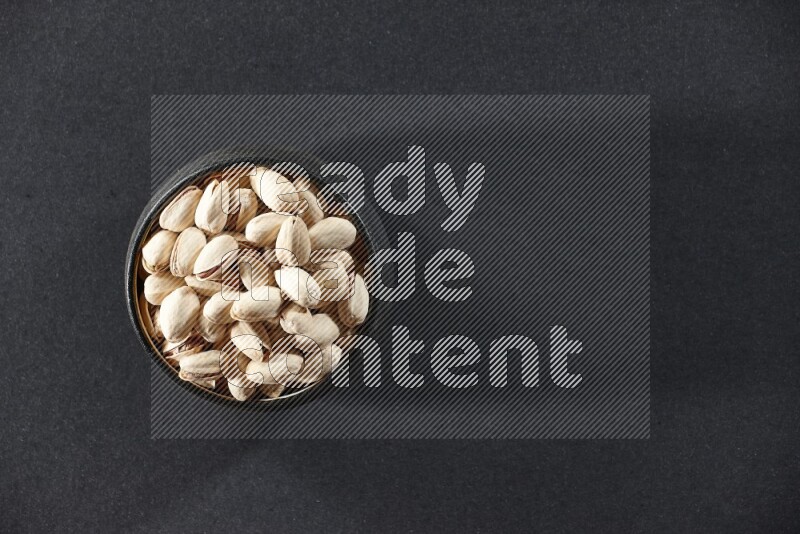 A black pottery bowl full of pistachios on a black background in different angles